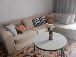 A beige sofa with striped and patterned cushions, a round white table with a vase of flowers, and a chevron-patterned rug. - Senere 1BR Apartment next to a premier mall (Nairobi)