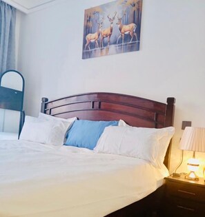 A bed with a wooden headboard, white and blue pillows, a nightstand with a lamp, and a painting of deer on the wall. - Senere 1BR Apartment next to a premier mall (Nairobi)