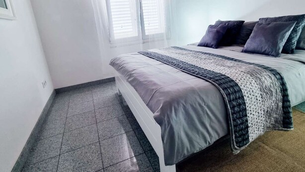 Ein Schlafzimmer mit einem Bett, einem Fenster mit weißen Vorhängen und einem Fliesenboden.