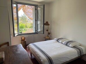 Ein Schlafzimmer mit Bett, Tisch, Lampe und einem Fenster mit geöffnetem Klappladen.