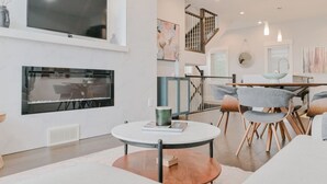 A modern living room with a flat-screen TV, fireplace, dining area, and staircase.