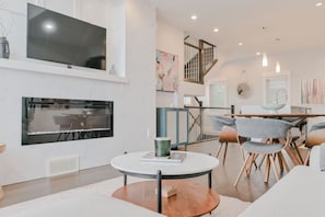A modern living room with a flat-screen TV, fireplace, dining area, and staircase. - 3BR | Garage | DT Views | Central | Lux Townhome (Calgary)