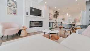 A modern living room with a flat-screen TV, a fireplace, a dining area with a staircase, and a large window.
