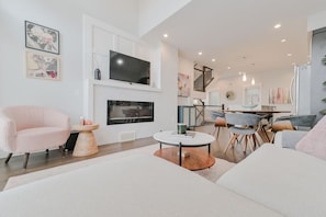 A modern living room with a flat-screen TV, a fireplace, a dining area with a staircase, and a large window. - 3BR | Garage | DT Views | Central | Lux Townhome (Calgary)