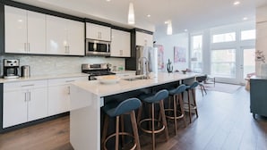 A modern kitchen with white cabinets, stainless steel appliances, a central island with stools, and large windows.