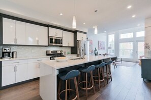 A modern kitchen with white cabinets, stainless steel appliances, a central island with stools, and large windows. - 3BR | Garage | DT Views | Central | Lux Townhome (Calgary)