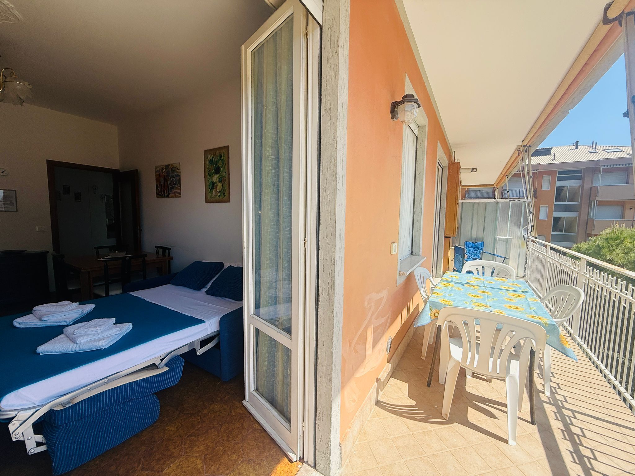 Un balcón con mesa y sillas, una cama con ropa de cama azul, y una puerta corrediza de vidrio.