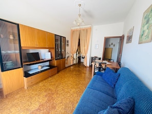 Ein Wohnzimmer mit blauem Sofa, Holzschränken, einem Fernseher und einem Essbereich mit Tisch und Stühlen.
