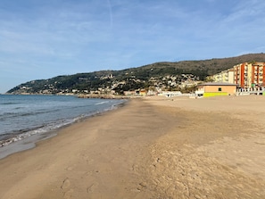 Ein Strand mit sandigem Ufer, ein Gebäude mit gelber Fassade und eine Hügelschaft mit Gebäuden im Hintergrund.