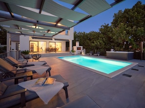 Ein moderner Poolbereich im Freien mit Liegestühlen, einem Essplatz und einer überdachten Terrasse.