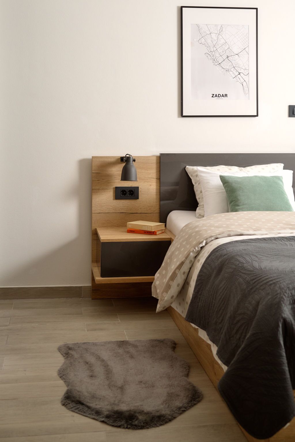A bedroom with a bed, a nightstand with a lamp, a framed map on the wall, and a rug on the floor.