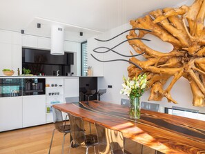 Une cuisine moderne avec des meubles blancs, une grande table à manger en bois et une sculpture en bois originale accrochée au mur.