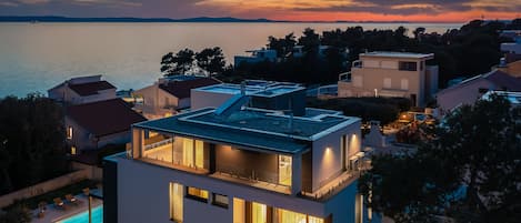 A modern two-story house with a rooftop terrace, a swimming pool, and a view of the sea at sunset.