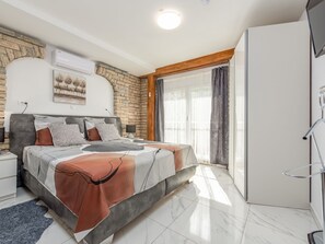 Une chambre à coucher comprenant un lit, une table de chevet, une commode, un mur en pierre, une fenêtre avec des rideaux et un luminaire au plafond.