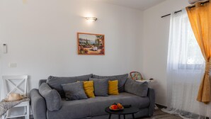 Ett vardagsrum med en grå soffa, ett litet bord med en skål med frukt, en tavla på väggen och ett fönster med gardiner.