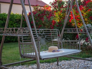 A metal swing chair with a cushion and a woven hat, set against a backdrop of red flowers and greenery. - Holiday Home Beti with heated pool by Interhome (Vrsi)