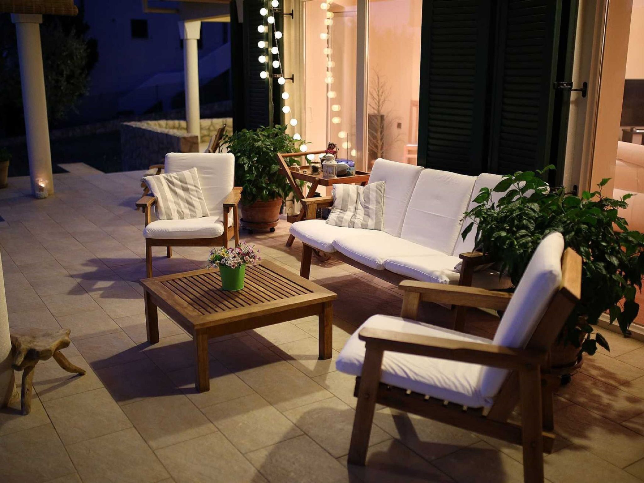 Un espace extérieur aménagé avec une table en bois, deux chaises blanches rembourrées et un canapé. On y trouve également des plantes en pot et une guirlande lumineuse.