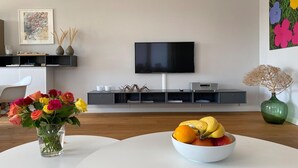 Un salon moderne avec un téléviseur à écran plat, une table à manger blanche agrémentée d’un panier de fruits et un vase rempli de fleurs.