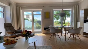 Un salon moderne comprenant un canapé, une table à manger et des chaises, un coin cuisine et de grandes fenêtres donnant sur un paysage verdoyant.