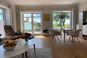 Ein modernes Wohnzimmer mit einer Couch, einem Esstisch und Stühlen, einer Küchenzeile sowie großen Fenstern mit Blick auf Grünflächen.