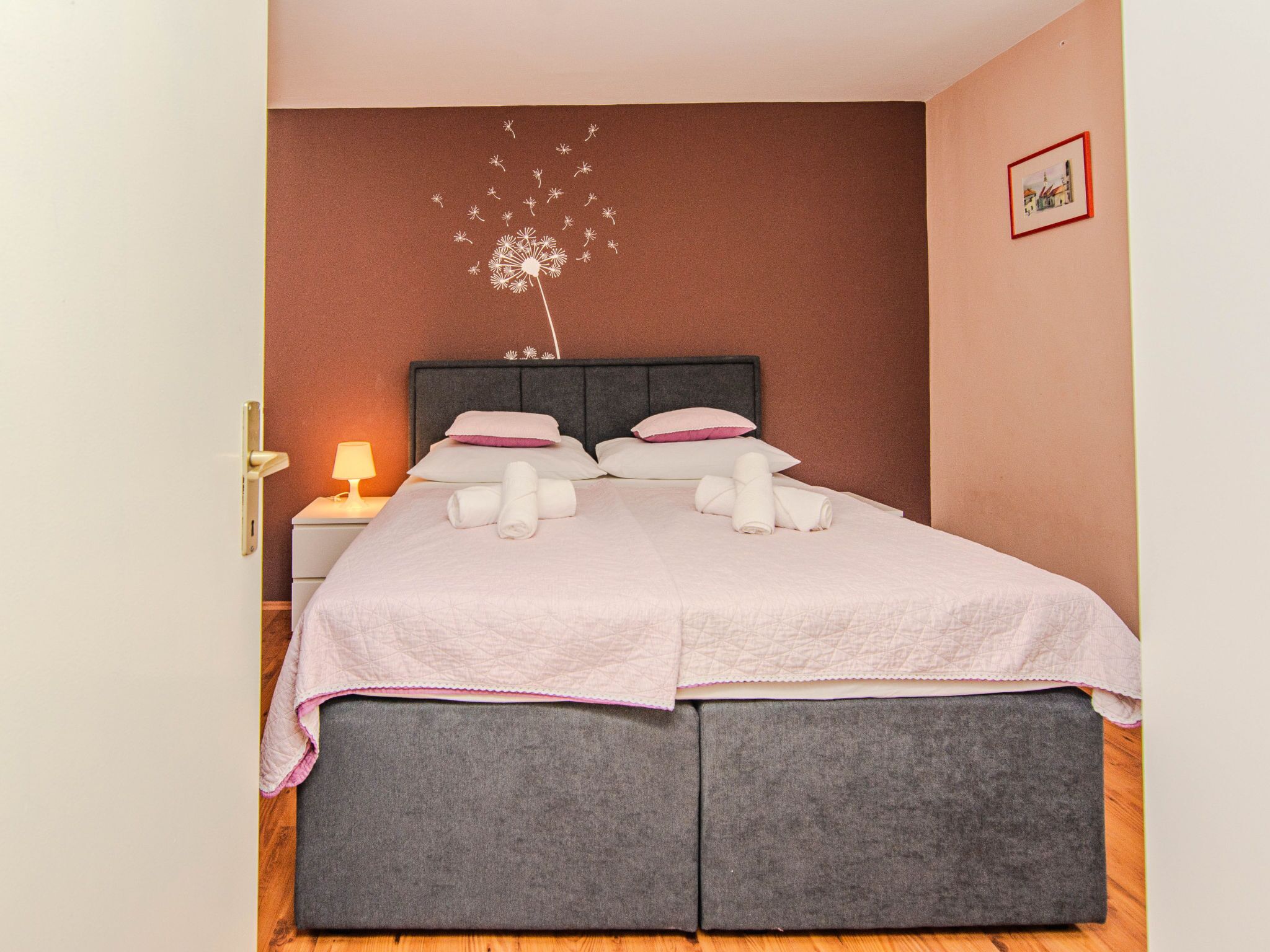 Ein Doppelbett mit grauem Kopfteil, Bettwäsche in Weiß und Rosa sowie ein Nachttisch mit Lampe.