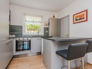 A compact kitchen with a built-in oven, a sink, and a window with a view of greenery. There are two bar stools and a framed picture on the wall. - Vacation home  by Interhome (Rovanjska)
