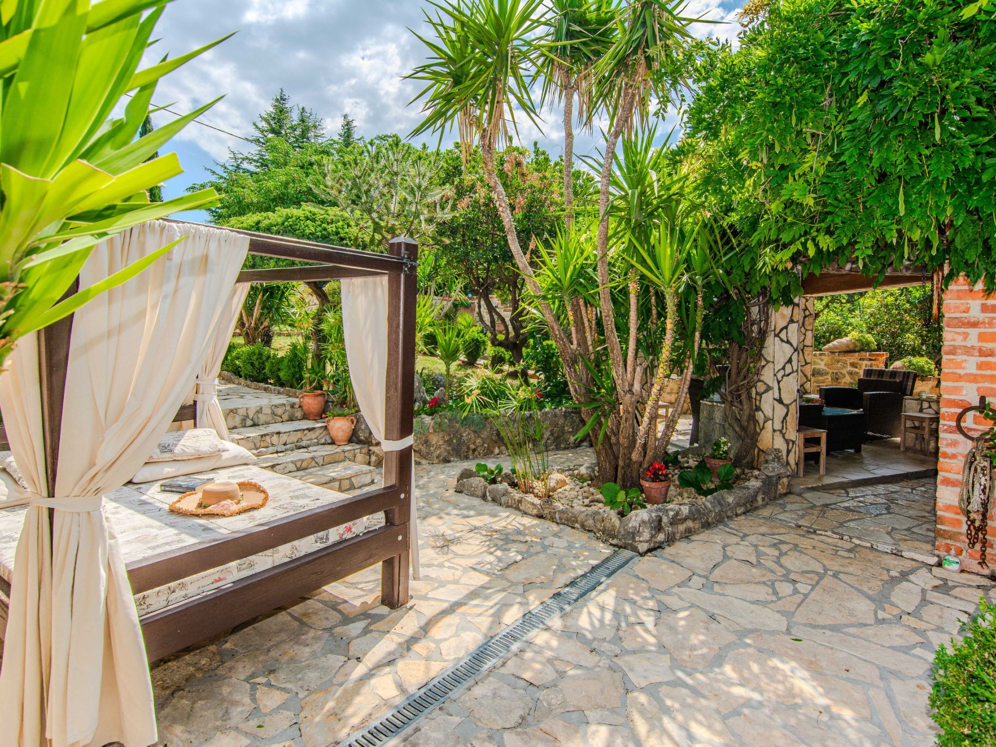 A stone patio with a bed and a dining area, surrounded by lush greenery and a brick wall.