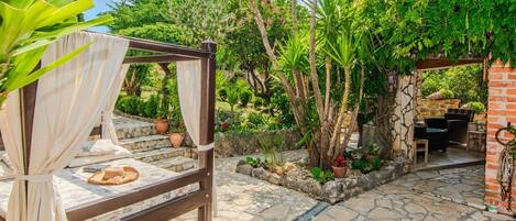 A stone patio with a bed and a dining area, surrounded by lush greenery and a brick wall.