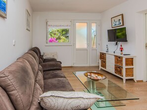 Una sala de estar con un sofá marrón, una mesa de centro de vidrio, un televisor en la pared y un ventanal con vistas a árboles y flores.