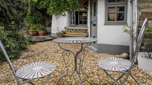A patio with two metal chairs and a table, a bench, and a house with a thatched roof and stone walls. - Host & Stay | Wistermead Cottage (Dolton)