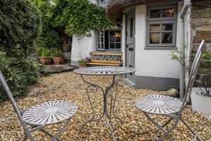 Eine Terrasse mit zwei Metallstühlen und einem Tisch, einer Bank sowie ein Haus mit einem Strohdach und Steinmauern.