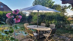 A round wooden table with four chairs and a large umbrella in a garden setting. - Ferienhaus in Püttsee auf Fehmarn (Püttsee)