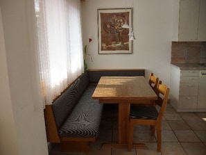A dining area with a wooden table and bench seating, a painting on the wall, and a kitchen area with cabinets. - Casa Migiur, (Laax Dorf), 37008 by Interhome (Laax)