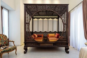 A traditional four-poster bed with ornate carvings, a floral-patterned armchair, and a small table with a lamp.