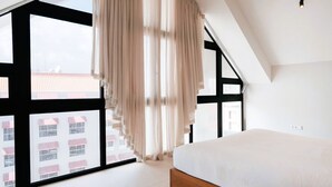 Quarto com uma janela ampla, uma cama e vista para prédios.