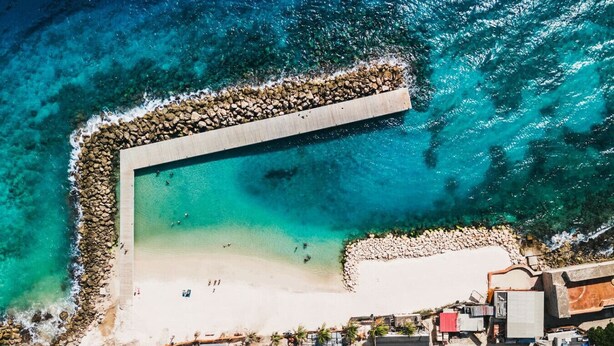 Vista aérea de uma praia com um molhe rochoso, águas turquesa cristalinas e uma faixa de areia.