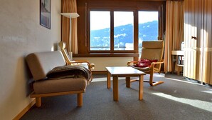 Een kamer met een bank, twee fauteuils, een bijzettafeltje, een raam met uitzicht op bergen en houten wandpanelen.