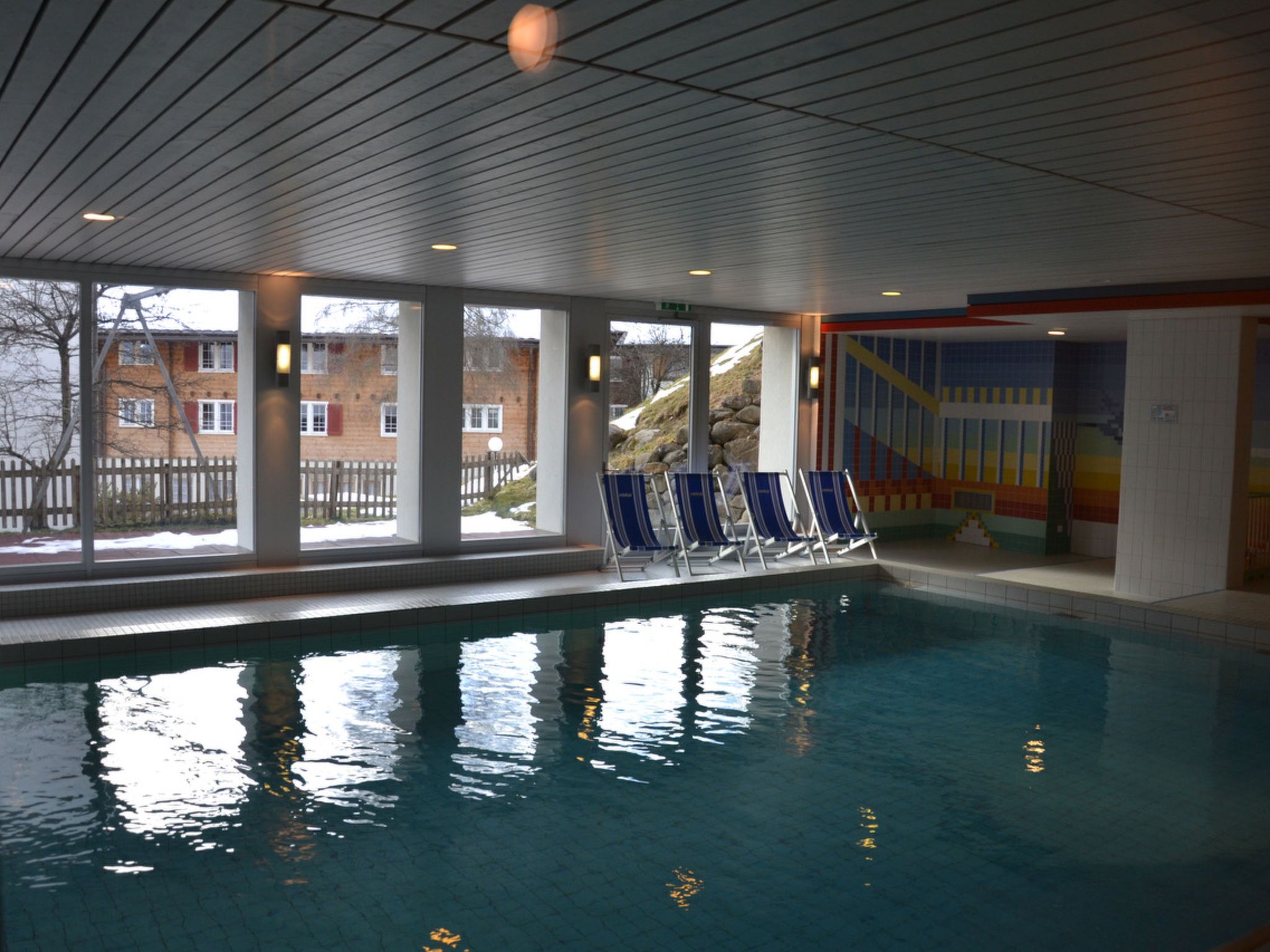 An indoor swimming pool with clear windows, blue lounge chairs, and a tiled ceiling.