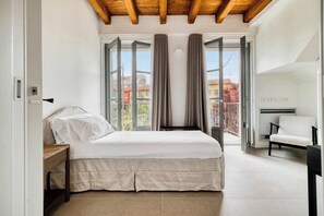 A bedroom with a bed, a nightstand, a chair, and a view of buildings through large windows. - w* | Luxury 5BR Retreat near Navigli (Milano)