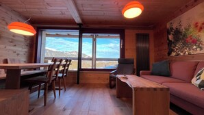 Un interior de madera con una mesa de comedor, sillas, un sofá y un gran ventanal con vistas a la montaña.