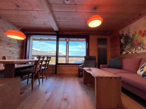 A wooden interior with a dining table, chairs, a sofa, and a large window offering a mountain view. - Ferienhaus Regan, Obersaxen-Valata, (Surcuolm), 38044B by Interhome (Surcuolm (Obersaxen))