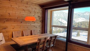 Un interior de madera con una mesa de comedor y sillas, una lámpara colgante, y un ventanal con vistas a la montaña.
