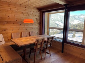 A wooden interior with a dining table and chairs, a hanging light fixture, and a large window with a mountain view. - Ferienhaus Regan, Obersaxen-Valata, (Surcuolm), 38044B by Interhome (Surcuolm (Obersaxen))