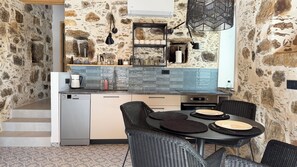 A compact kitchen with stone walls, a built-in oven, and a dining area with wicker chairs. - ELAFONISI PETRA HOUSE - Traditional Stone House near Elafonisi Beach 2BD, 2BTH (Kissamos, Chania)