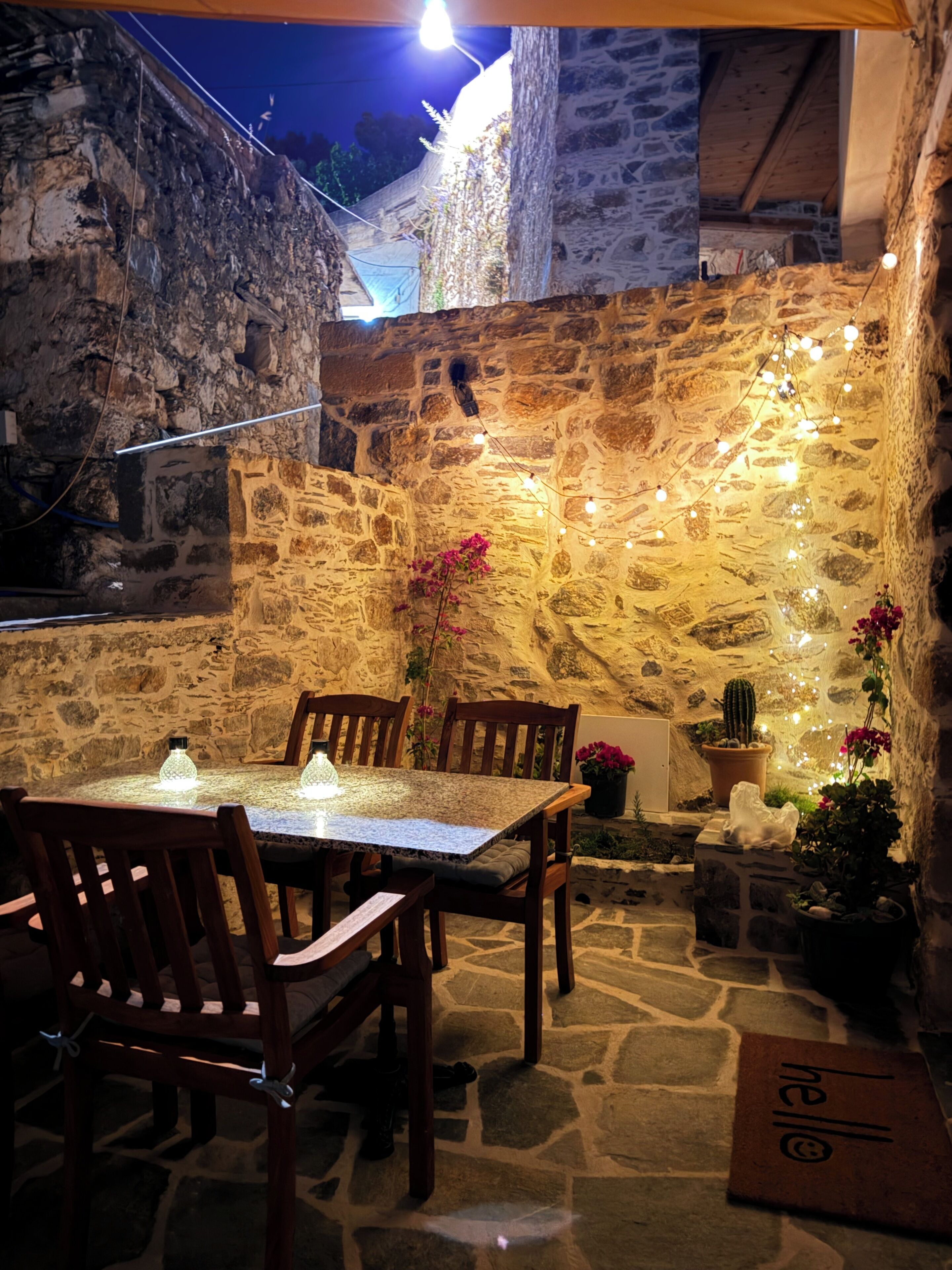 Un espace de restauration en plein air avec des murs en pierre, des chaises en bois, une table avec un plateau en verre, des guirlandes lumineuses et des plantes en pot.
