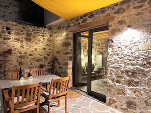 A stone-walled patio with a table and chairs, a glass door leading inside, and string lights. - ELAFONISI PETRA HOUSE - Traditional Stone House near Elafonisi Beach 2BD, 2BTH (Kissamos, Chania)