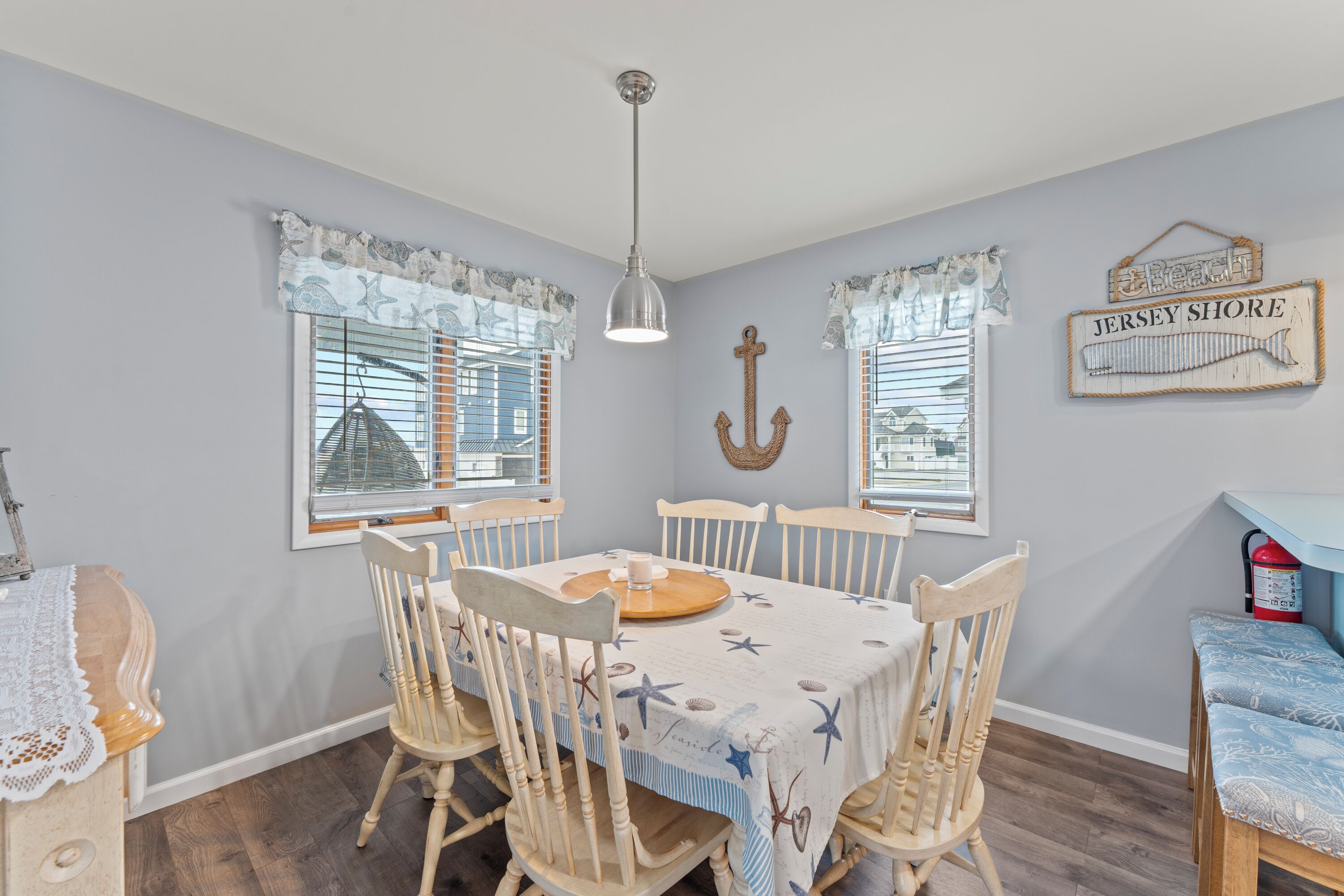 A dining area with a table set for four, surrounded by wooden chairs, a hanging light fixture, and nautical-themed window treatments.
