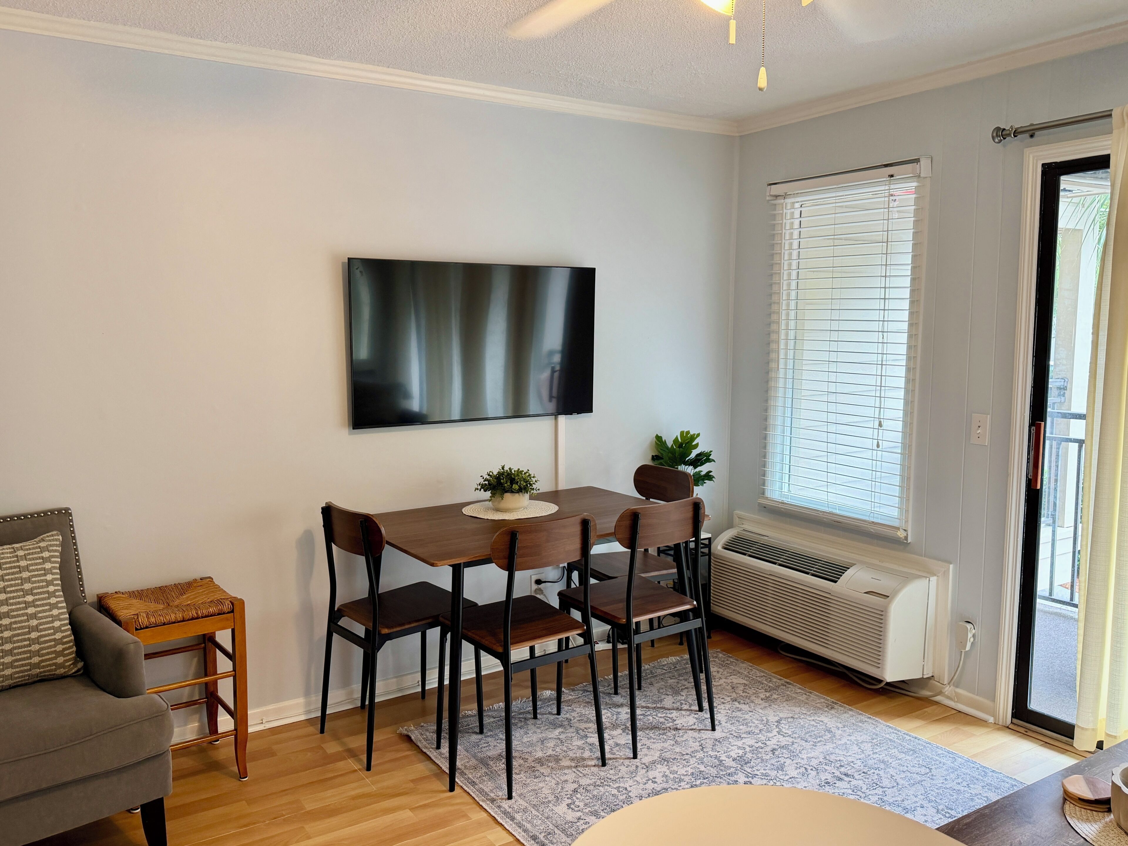Un coin repas comprenant une table, des chaises, un téléviseur à écran plat, une petite plante et une fenêtre avec des stores.