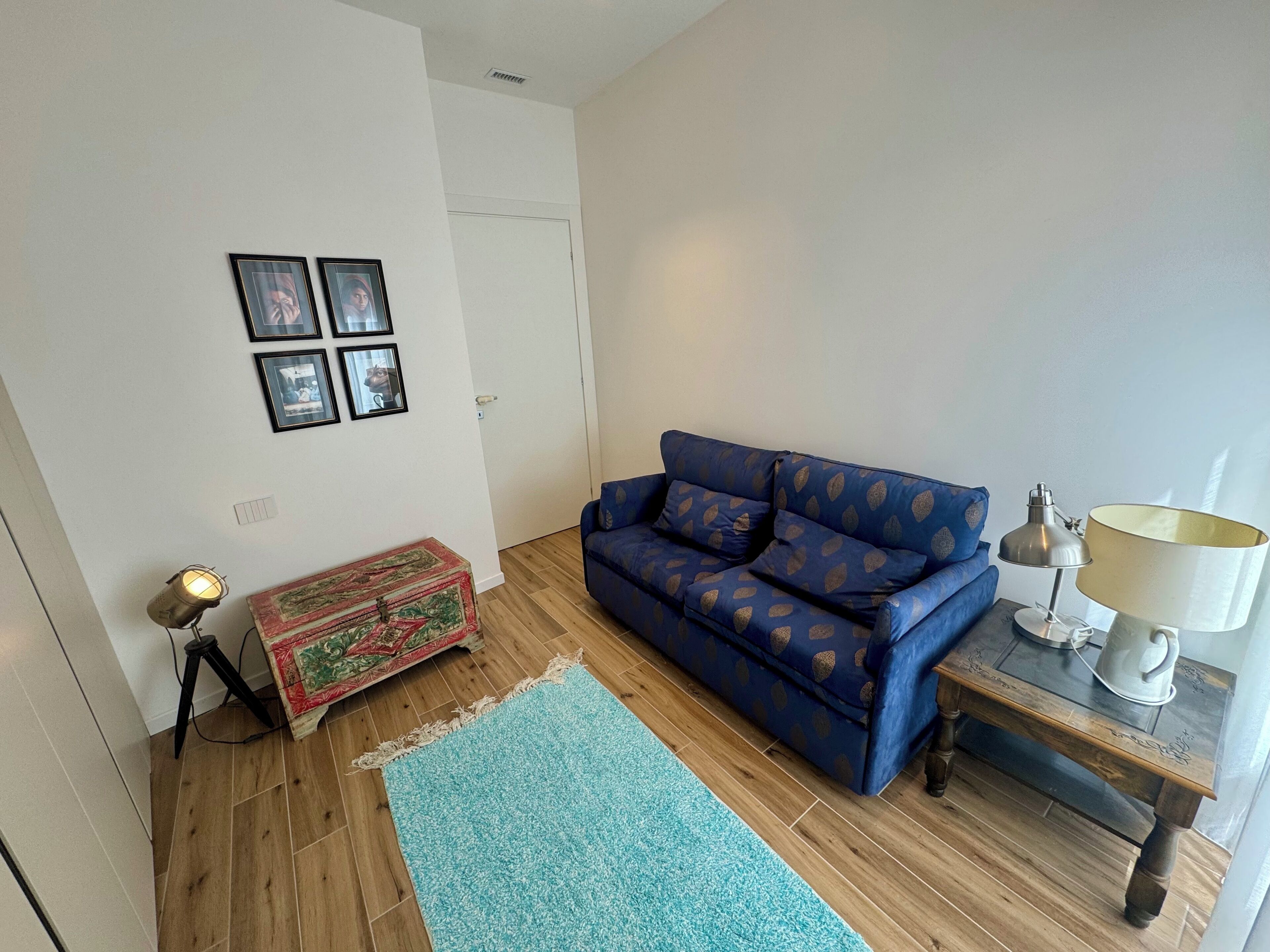 Un salon avec un canapé bleu, une petite table avec une lampe, un plancher en bois et un tapis à motifs.