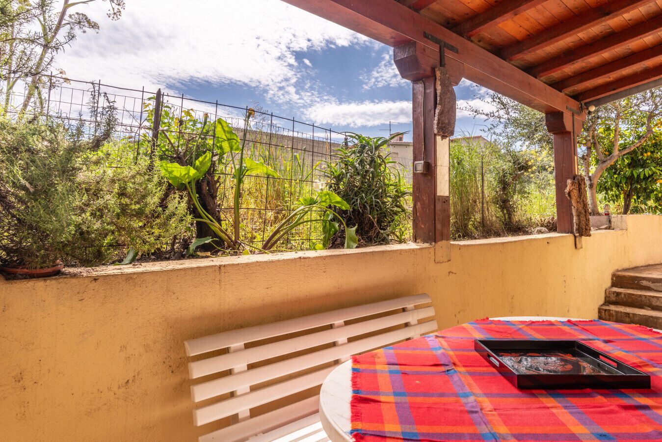 A covered outdoor area with a table, a bench, and a view of greenery and a fence.